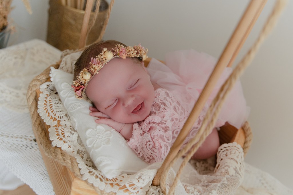 Bébé souriant endormi dans un panier en osier, portant une robe en dentelle rose et un diadème floral.