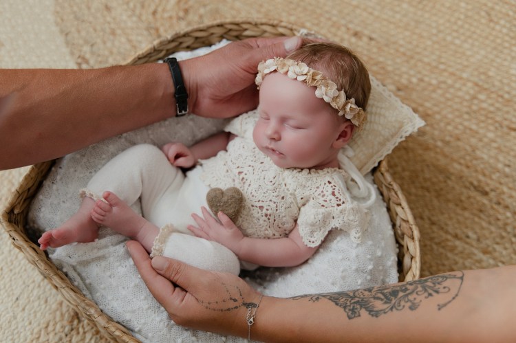 Un bébé endormi dans un panier, portant un vêtement en crochet et une couronne de fleurs, avec des mains qui le soutiennent délicatement.